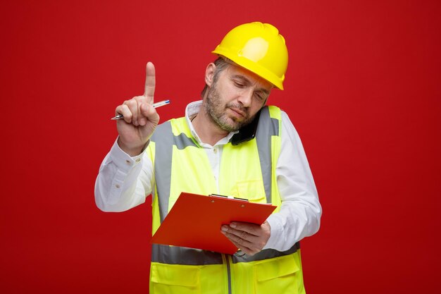Мужчина-строитель в строительной форме и защитном шлеме держит буфер обмена, разговаривает по мобильному телефону, выглядит недовольным и просит подождать, показывая указательный палец, стоящий на красном фоне