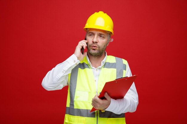 Мужчина-строитель в строительной форме и защитном шлеме держит буфер обмена и разговаривает по мобильному телефону, выглядя недовольным, кривя рот, стоя на красном фоне