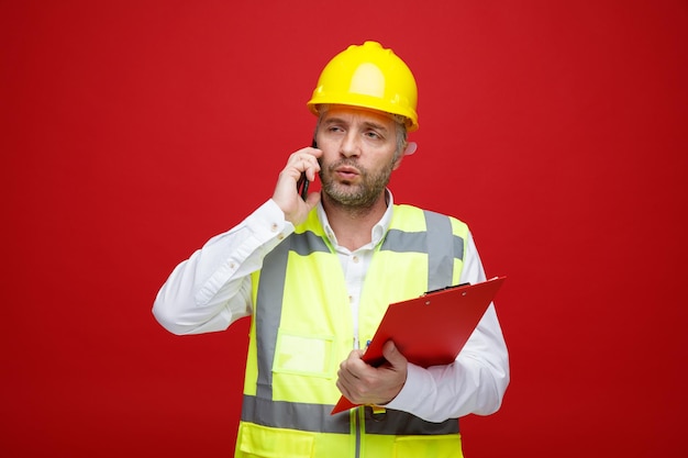 Builder man in construction uniform and safety helmet holding clipboard talking on mobile phone looking displeased making wry mouth standing over red background