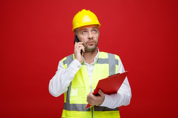 Builder man in construction uniform and safety helmet holding clipboard talking on mobile phone looking up puzzled standing over red background