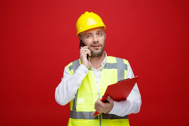 건설 유니폼을 입은 빌더 남자와 클립보드를 들고 있는 안전 헬멧이 빨간색 배경 위에 서서 회의적인 표정으로 웃고 있는 휴대전화에 대해 이야기하고 있다