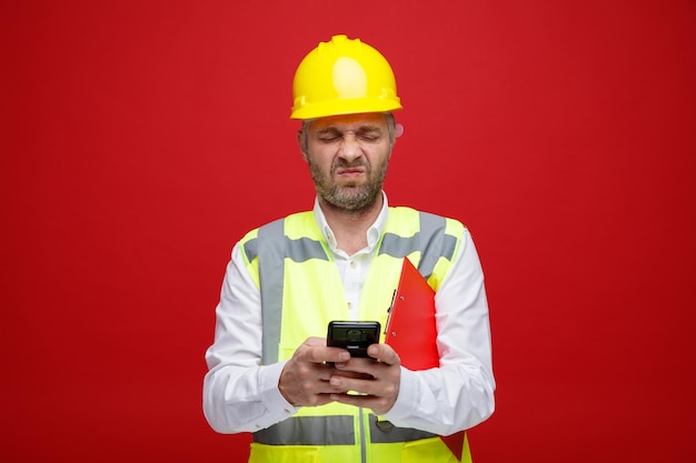 Builder man in construction uniform and safety helmet holding clipboard texting message using smartphone frowning making wry mouth being dissatisfied standing over red background