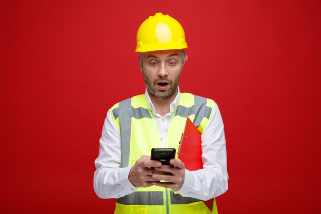 Мужчина-строитель в строительной форме и защитном шлеме держит смс-сообщение в буфере обмена с помощью смартфона, выглядя изумленным и удивленным, стоя на красном фоне
