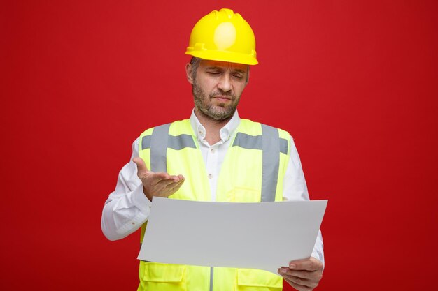 Builder man in construction uniform and safety helmet holding a plan looking at it being confused raising arm in displeasure standing over red background