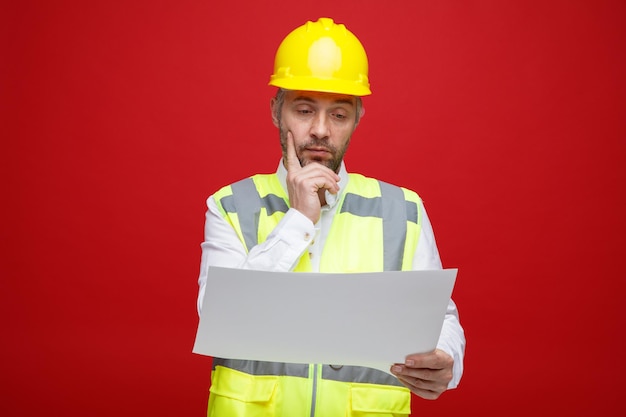 Builder man in construction uniform and safety helmet holding a plan looking at it with interest thinking standing over red background