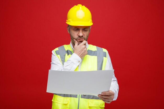 Builder man in construction uniform and safety helmet holding a plan looking at it with pensive expression thinking with hand on his chin standing over red background