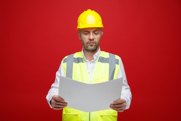 赤い背景の上に立っている真面目な顔でそれを見て計画を保持している建設制服と安全ヘルメットのビルダーの男