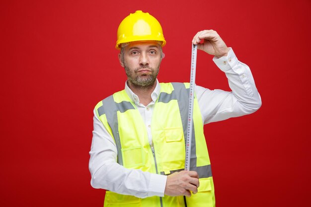 Builder man in construction uniform and safety helmet holding ruler looking at camera with serious face standing over red background