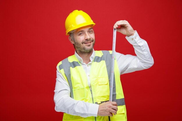 Builder man in construction uniform and safety helmet holding ruler looking at camera with smile on face standing over red background