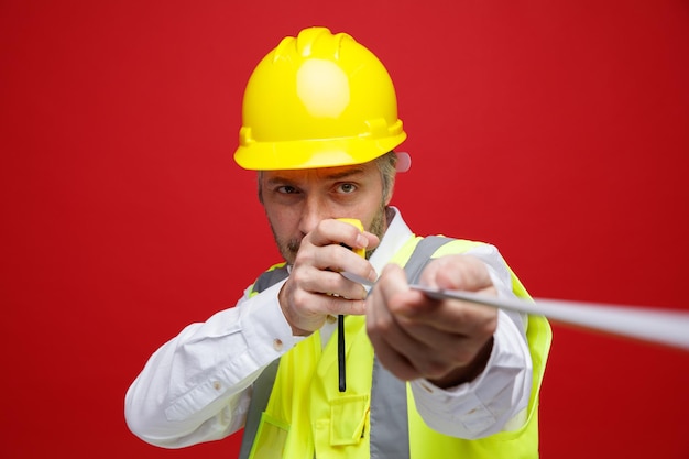 Builder man in construction uniform and safety helmet holding ruler pointing it at camera looking confident standing over red background