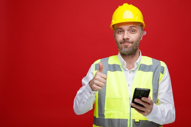 赤い背景の上に立って親指を示す幸せでポジティブな笑顔のカメラを見てスマートフォンを保持している建設制服と安全ヘルメットのビルダーの男