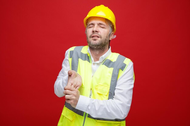 빨간색 배경 위에 서서 손을 쭉 뻗고 피곤하고 과로해 보이는 안전 헬멧을 쓴 건설업자 남자