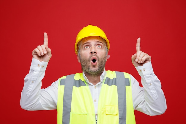 Uomo del costruttore in uniforme da costruzione e casco di sicurezza che osserva in su sorpreso puntando con il dito indice in piedi su sfondo rosso