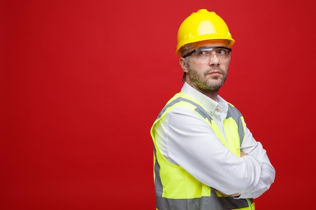 Free photo builder man in construction uniform and safety helmet wearing safety glasses looking aside with pensive expression with arms crossed on his chest standing over pink background