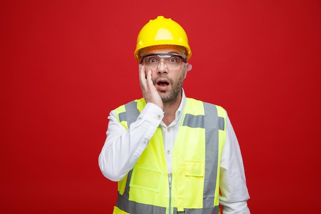 Мужчина-строитель в строительной форме и защитном шлеме в защитных очках смотрит в камеру, пораженный и удивленный, держа руку на щеке, стоя на красном фоне