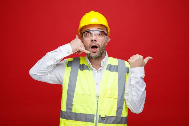 Мужчина-строитель в строительной форме и защитном шлеме в защитных очках смотрит в камеру, пораженный и удивленный, делая жест "позвони мне", указывая большим пальцем в сторону, стоя на красном фоне