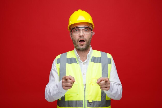 Строитель в строительной форме и защитном шлеме в защитных очках смотрит в камеру, пораженный и удивленный, указывая указательным пальцем на камеру, стоящую на розовом фоне