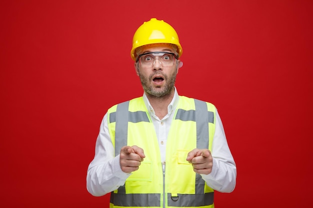 Builder man in construction uniform and safety helmet wearing safety glasses looking at camera amazed and surprised pointing with index fingers at camera standing over pink background