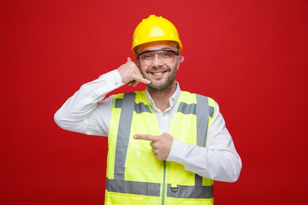 Builder man in construction uniform and safety helmet wearing safety glasses looking at camera smiling cheerfully making call me gesture pointing with index finger to the side standing red background