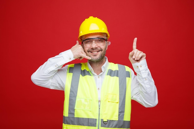Builder man in construction uniform and safety helmet wearing safety glasses looking at camera smiling cheerfully making call me gesture pointing with index finger up standing over red background