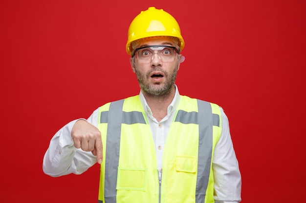 건설 유니폼을 입은 건축업자 남자와 안전 안경을 쓴 안전 헬멧이 분홍색 배경 위에 서 있는 검지 손가락으로 가리키는 카메라를 보고 놀란다