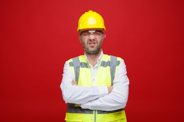 건설 유니폼과 안전 헬멧을 쓴 빌더 남자는 분홍색 배경 위에 서 있는 가슴에 팔짱을 끼고 역겨운 표정으로 카메라를 바라보고 있다