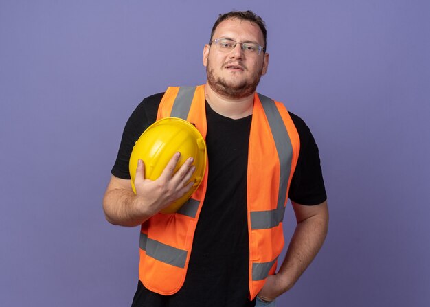 Builder man in construction vest holding safety helmet looking at camera smiling confident