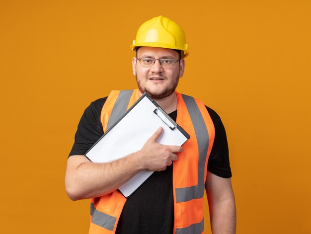 Builder man in construction vest and safety helmet holding clipboard looking at camera