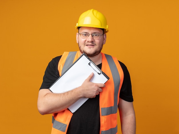 Человек-строитель в строительном жилете и защитном шлеме с буфером обмена, смотрящим в камеру