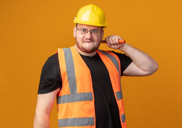 Builder man in construction vest and safety helmet holding hammer looking at camera with angry face