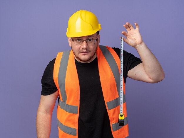 Builder man in construction vest and safety helmet holding measure tape looking at camera surprised