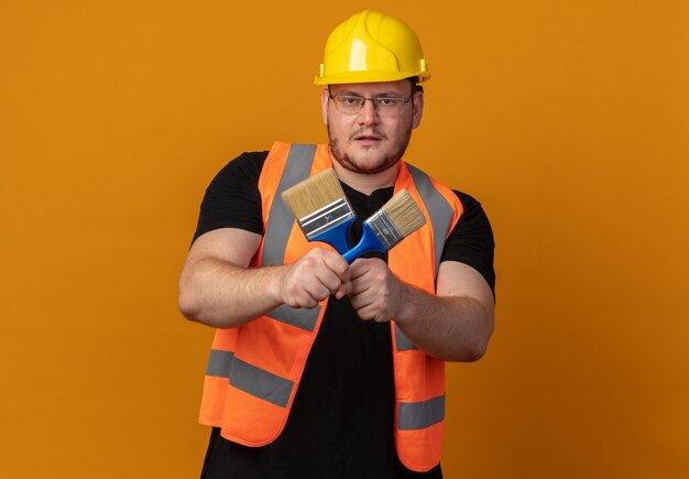 Builder man in construction vest and safety helmet holding paint brushes looking at camera with confident expression