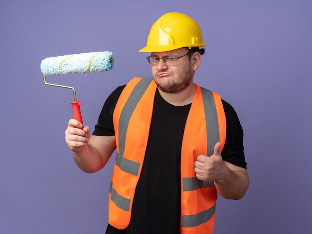 Builder man in construction vest and safety helmet holding paint roller looking at camera showing thumbs up standing over blue background