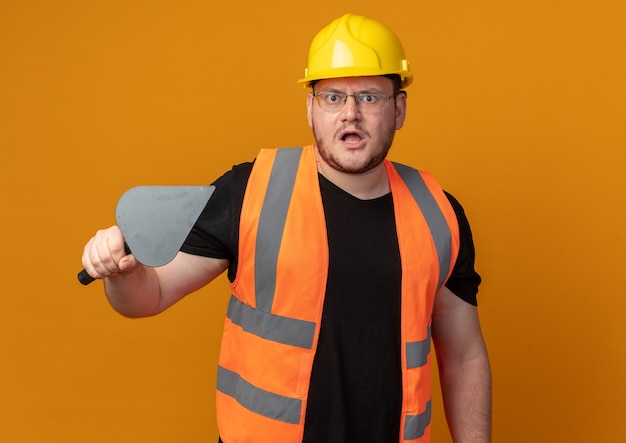 Uomo del costruttore in giubbotto da costruzione e casco di sicurezza che tiene una spatola guardando la telecamera dispiaciuta con la faccia arrabbiata in piedi su sfondo arancione