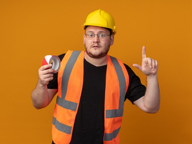 건설 조끼를 입은 빌더 남자와 검지 손가락으로 가리키는 스카치 테이프를 들고 주황색 배경 위에 서 있는 걱정스러운 표정