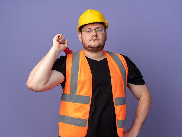 Человек-строитель в строительном жилете и защитном шлеме с гаечным ключом смотрит в камеру с серьезным уверенным выражением лица, стоящим над синим