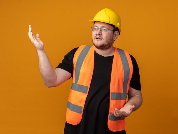 Builder man in construction vest and safety helmet looking aside confused with arms raised
