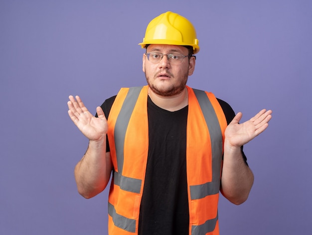 Человек-строитель в строительном жилете и защитном шлеме, смотрящий в камеру, сбивает с толку, разводя руки в стороны, стоя над синим
