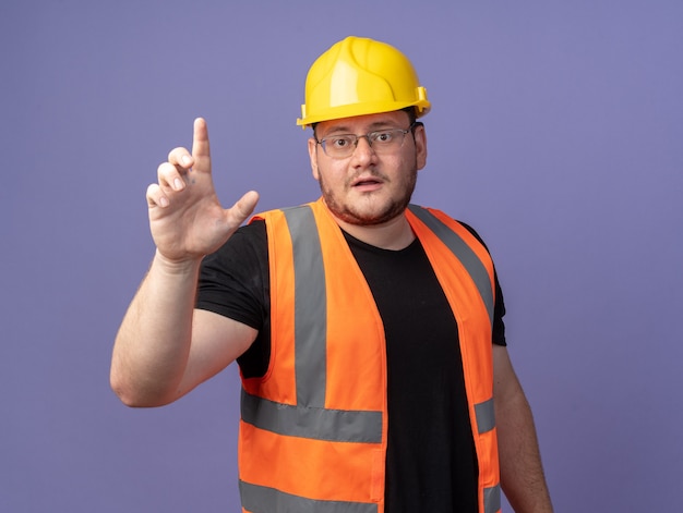 Человек-строитель в строительном жилете и защитном шлеме смотрит в камеру, показывая предупреждение указательным пальцем, стоящее на синем фоне