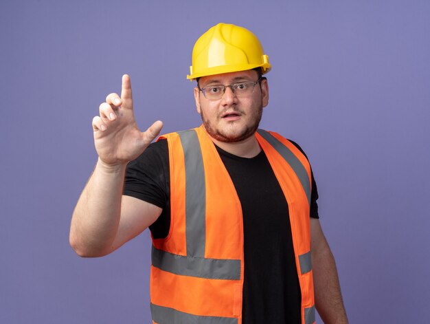 Builder man in construction vest and safety helmet looking at camera showing index finger warning standing over blue background