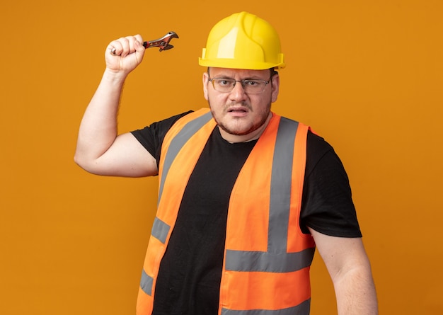 Builder man in construction vest and safety helmet looking at camera with angry face swinging a wrench standing over orange