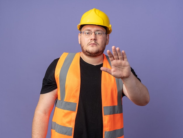 Человек-строитель в строительном жилете и защитном шлеме смотрит в камеру с серьезным лицом, делая стоп-жест рукой, стоящей над синим