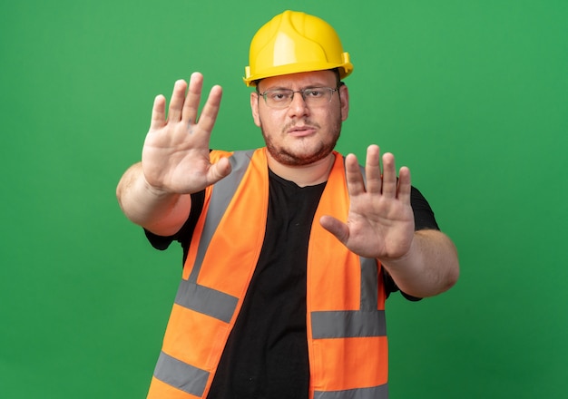 Человек-строитель в строительном жилете и защитном шлеме смотрит в камеру с серьезным лицом, делая жест стоп с руками, стоящими над зеленым