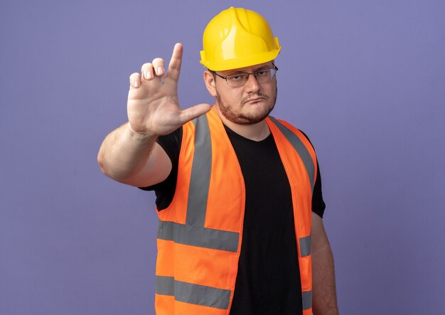 Builder man in construction vest and safety helmet looking at camera with serious face showing index finger warning gesture standing over blue