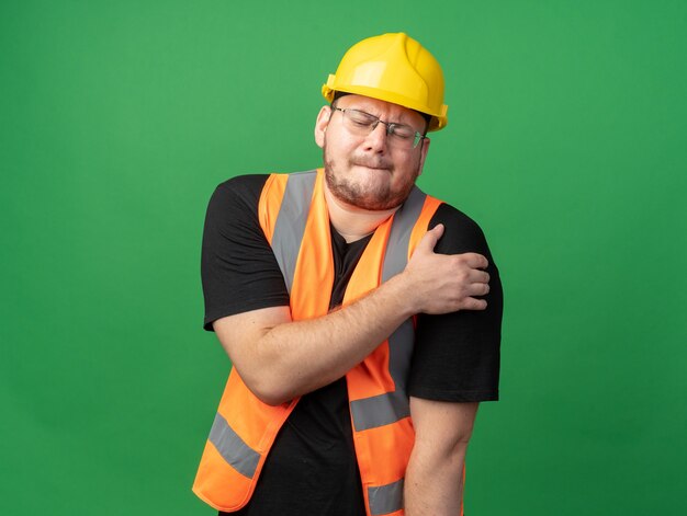 Builder man in construction vest and safety helmet looking unwell touching his shoulder feeling pain standing over green