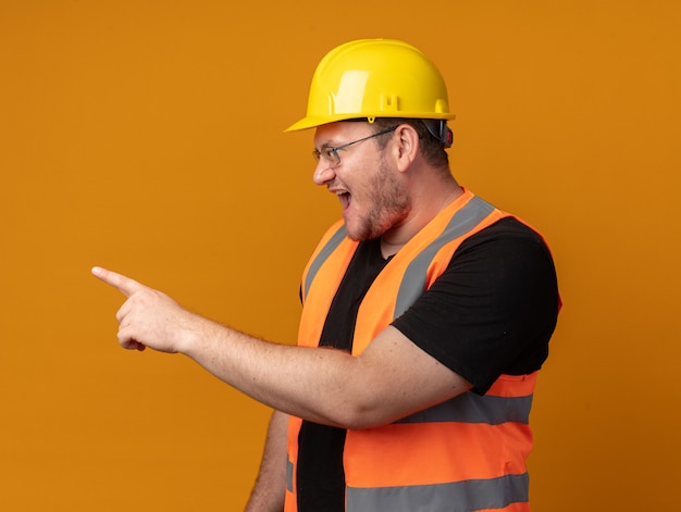 オレンジ色の背景の上に立っている攻撃的な表情で叫んで人差し指で横を指している建設ベストと安全ヘルメットのビルダーの男