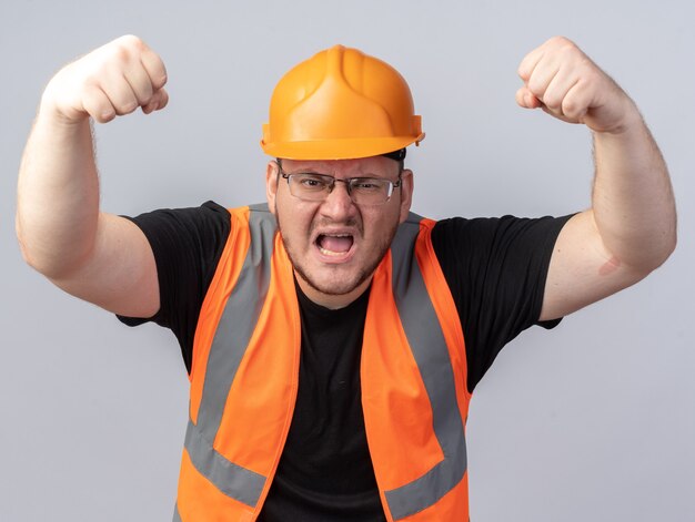 Человек-строитель в строительном жилете и защитном шлеме кричит с сердитым лицом, поднимая кулаки, стоя над белой