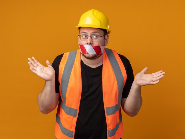 Builder man in construction vest and safety helmet with tape over his mouth looking confused spreading arms to the sides