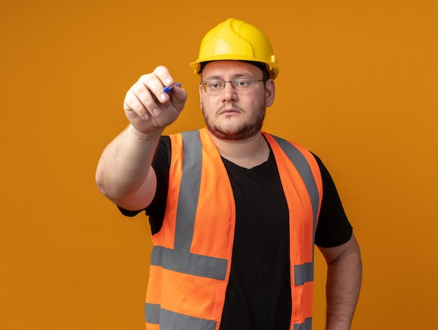 Человек-строитель в строительном жилете и защитном шлеме пишет ручкой что-то с серьезным лицом, стоящим на оранжевом фоне