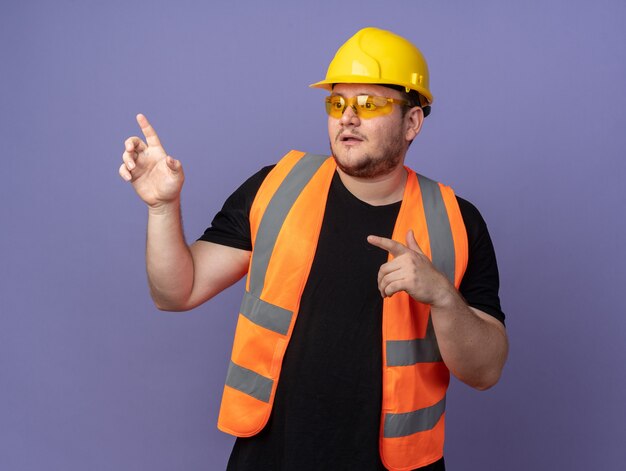 Builder man in construction vest yellow safety glasses and safety helmet looking confused pointing with index fingers at something